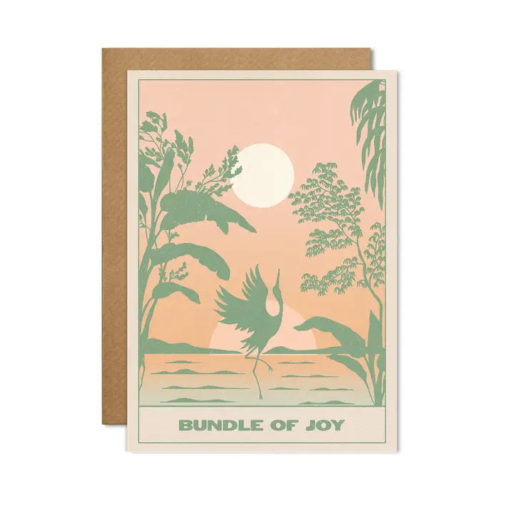 A greetings card with a light green frame around the side and an image of a pale pink and orange moonrise/sunset in the background and a sage green stork and foliage in the foreground. The words &#39;bundle of joy&#39; feature in sage green at the bottom.