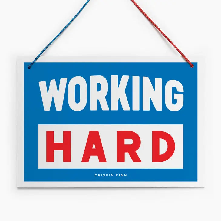 An image of a blue hanging sign with a vintage feel. It has the words 'working hard' in block capitals and a half red/half blue hanging cord.