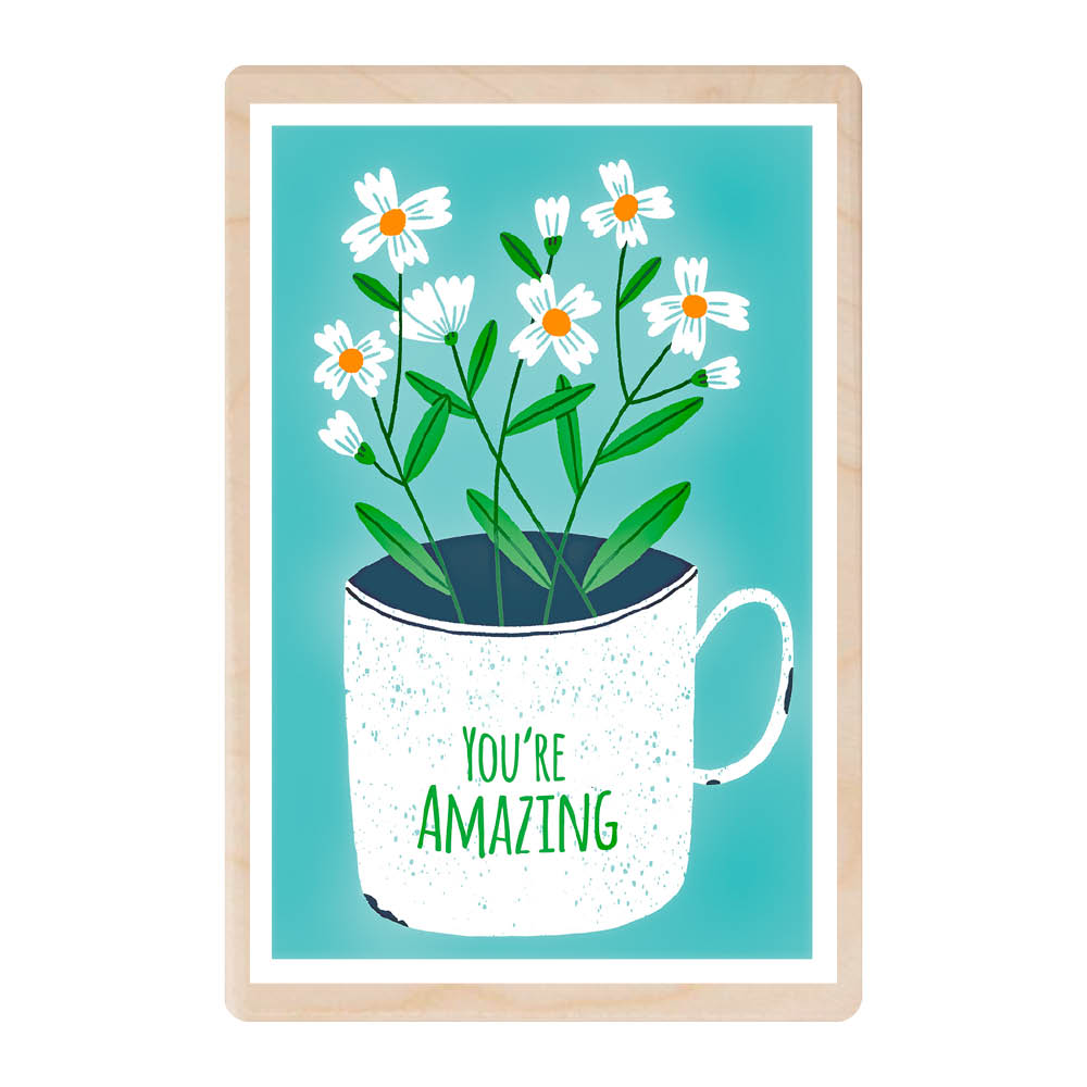A wooden postcard featuring a mug with the words &#39;you&#39;re amazing&#39; on the side with daisy flowers arranged in it. The mug is white and the letters are green..