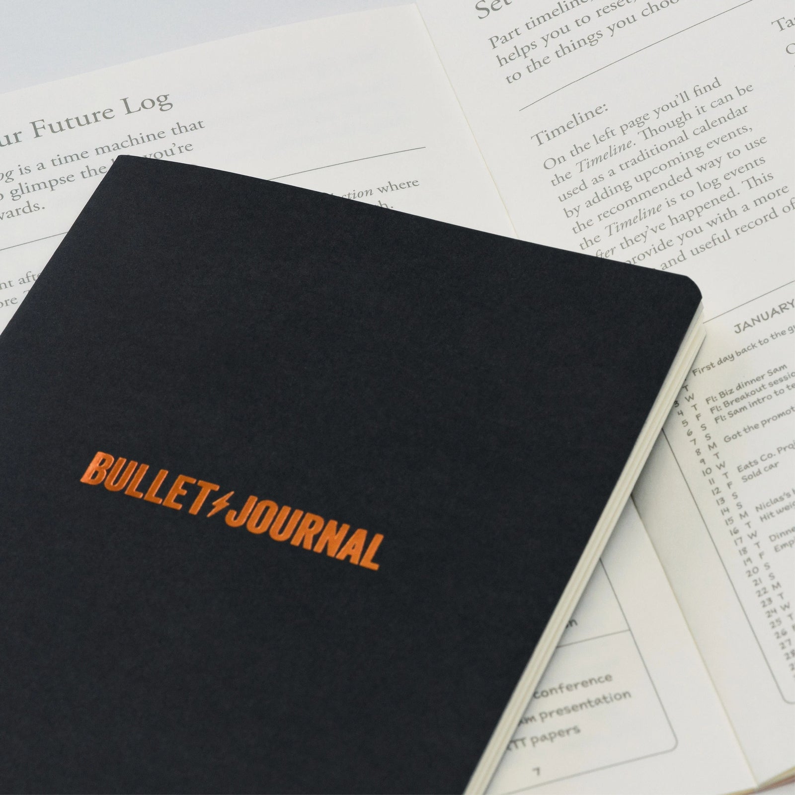 An image of a black notebook with bronze block writing saying 'bullet journal' with a lightening bolt between the words. It is sitting across typed instructions on white paper.