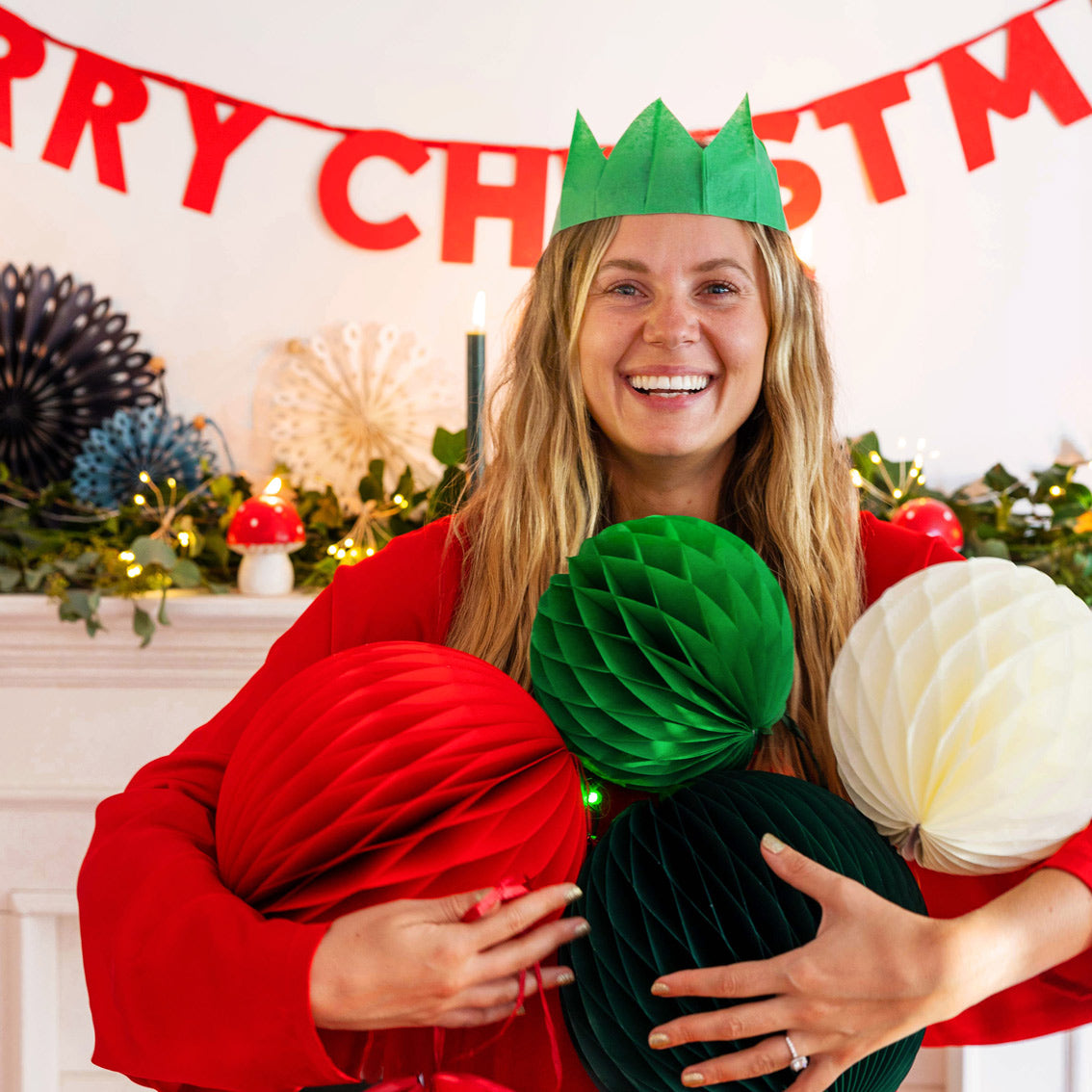 An image of a white woman with blonde hair holding 4 christmas paper honeycomb ball decorations in her arms. She has a green cracker party hat on, a red jumper and is standing infront of a festively decorated fireplace.
