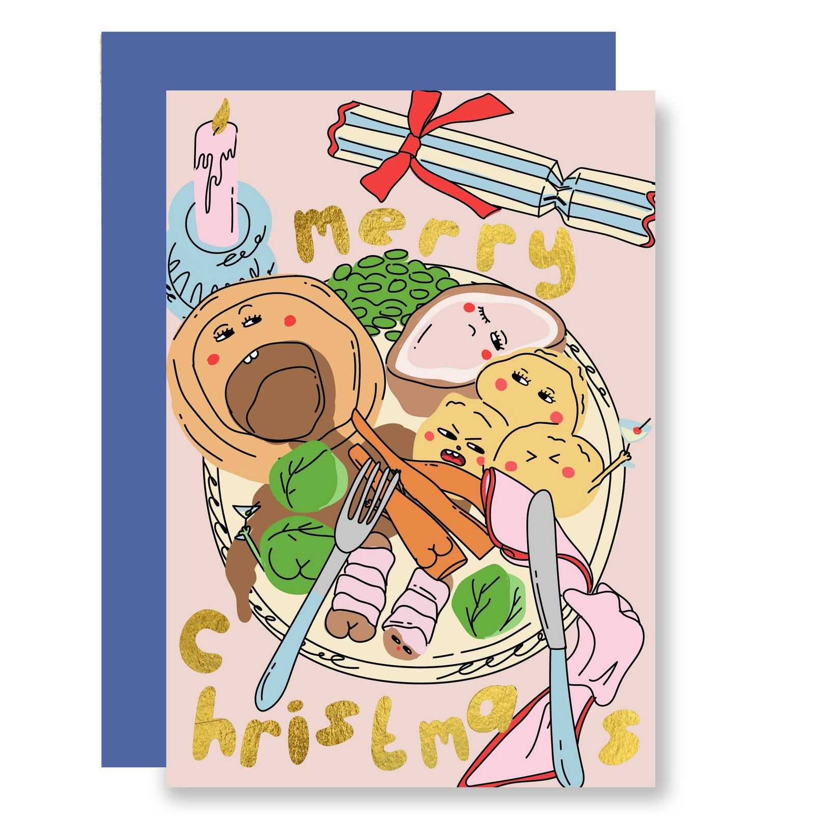 A Christmas greeting card with a light pink background and a blue envelope behind it. The card features a whimsical illustration of a Christmas feast on a plate with potatoes, brussel sprouts, a candle, crackers, and the words 'merry Christmas' in gold lettering.