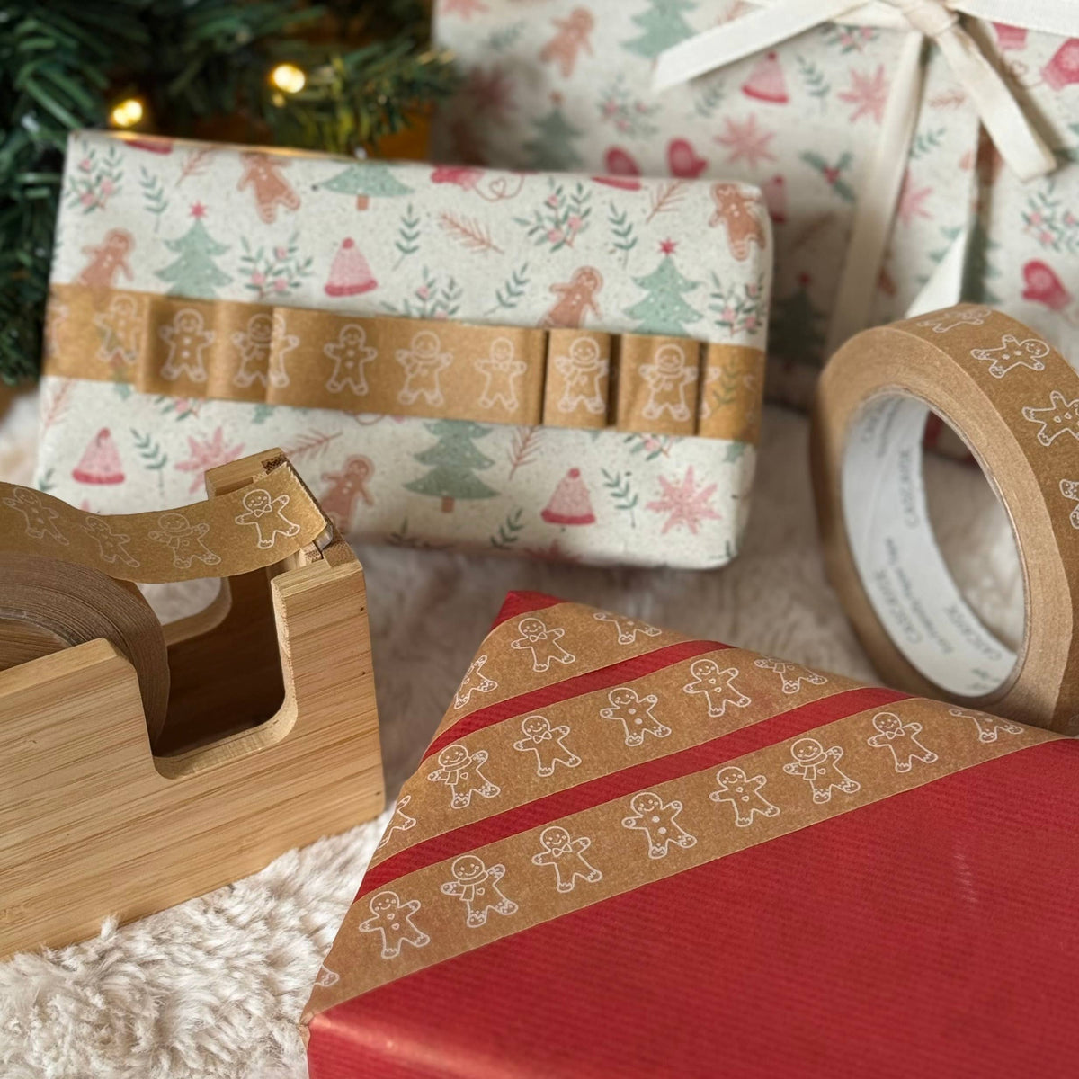Christmas-themed gift wrapping supplies including patterned wrapping paper, brown washi tape with gingerbread men, a wooden tape dispenser, and a tape roll on a white faux fur surface with wrapped presents and a Christmas tree in the background.