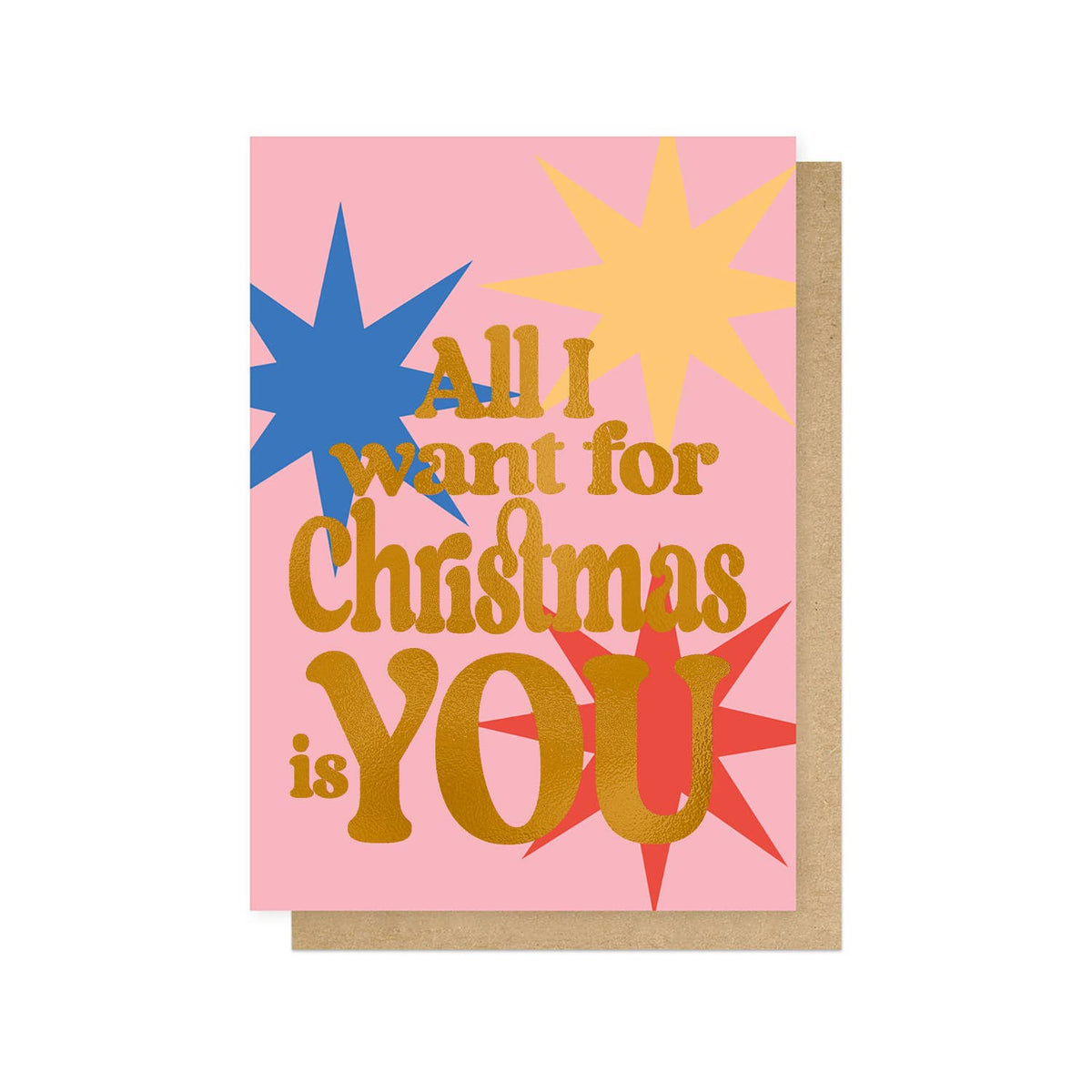 A pink greeting card with gold foil text reading 'All I want for Christmas is YOU' and colourful starburst graphics in blue, yellow, red, and orange, shown with a brown kraft envelope on a white background.