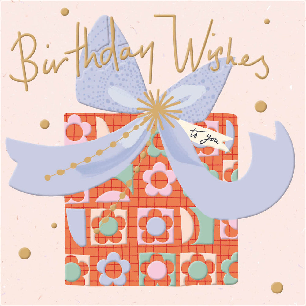 A greetings card with a light pink background, featuring a large square wrapped gift as the main image. It has a retro-style orange and floral wrapping paper and a large light blue bow on top. The words &#39;birthday wishes&#39; are written along the top in gold foil.