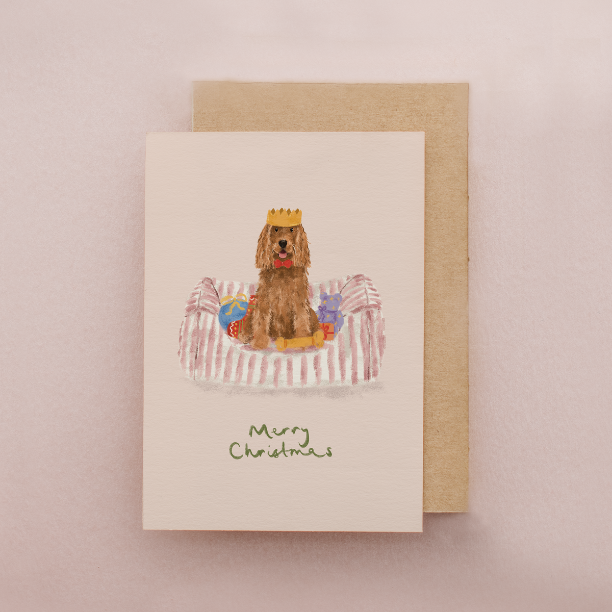 A Christmas greeting card featuring an illustration of a brown dog wearing a gold crown and sitting on a striped bed, with 'Merry Christmas' written below. The card is shown with a kraft envelope on a soft pink background.