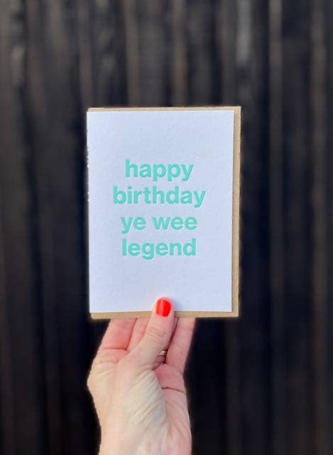 A hand with red nail polish holds an A6 letterpress printed birthday card that reads 'happy birthday ye wee legend' in turquoise text on a white background, set against a dark wood plank backdrop.
