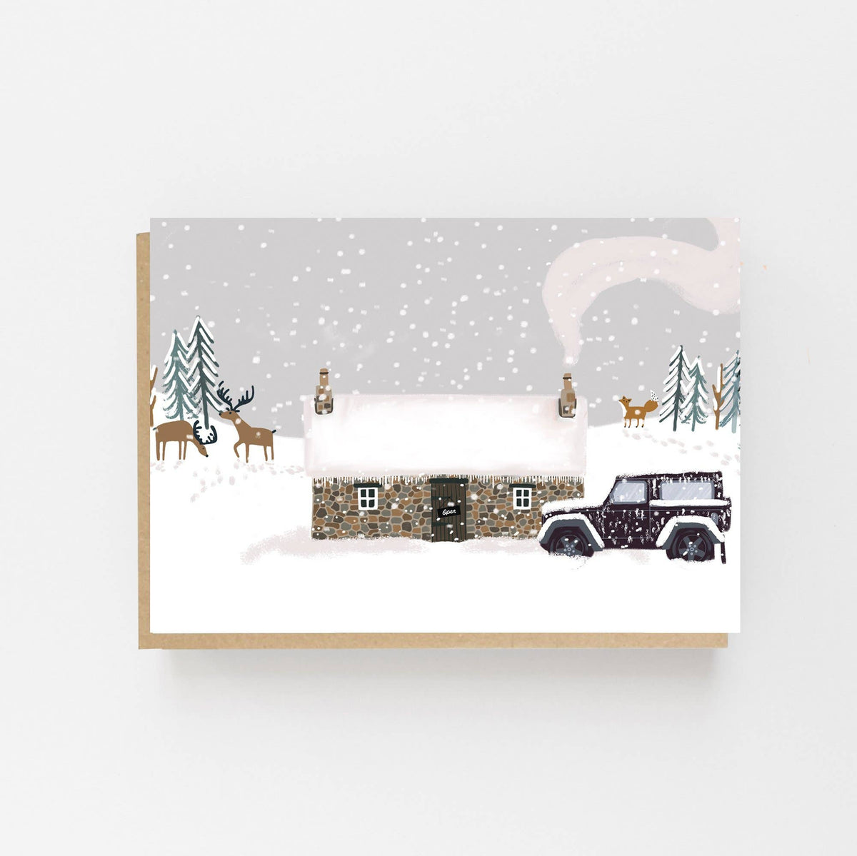 An A6 greeting card showing a snowy winter cottage bothy scene with pine trees, deer, a smoking chimney, and a dark land rover parked in the snow, displayed against a light gray background with a kraft envelope behind it.