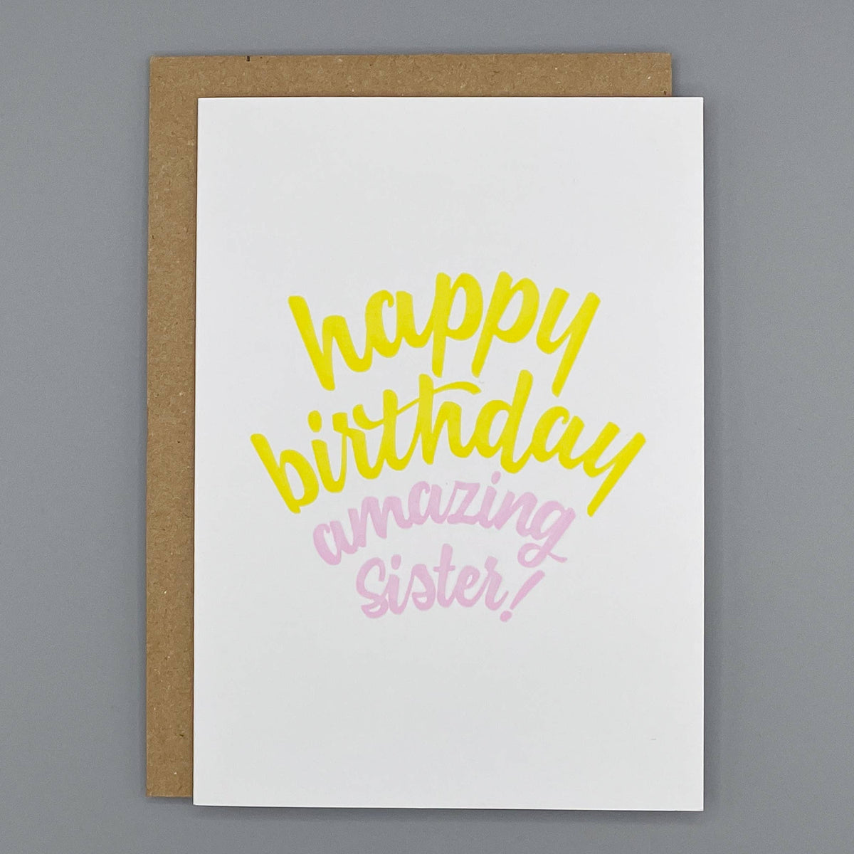 A cream letterpress printed greeting card with the words 'happy birthday amazing Sister!' in yellow and pink script, shown with a brown kraft envelope on a gray background.
