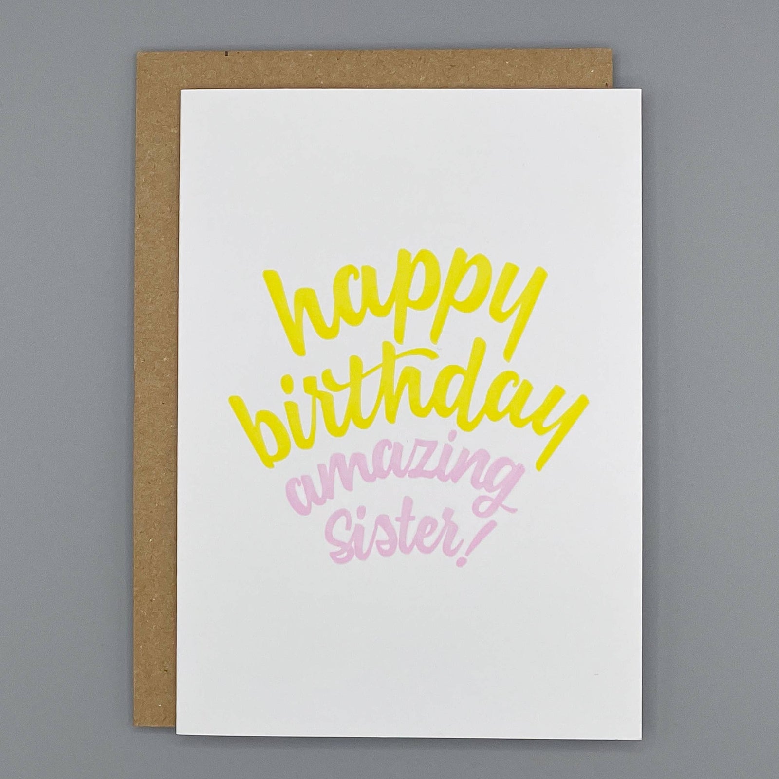 A cream letterpress printed greeting card with the words 'happy birthday amazing Sister!' in yellow and pink script, shown with a brown kraft envelope on a gray background.