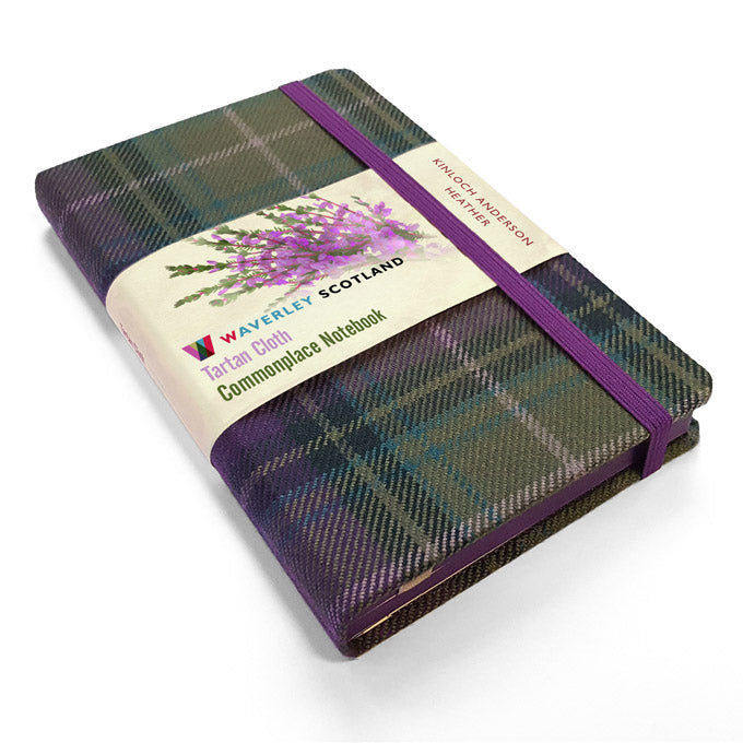 An image of a dark green and purple tartan notebook. It has a purple closure band and a paper belly band explaining the product and brand.