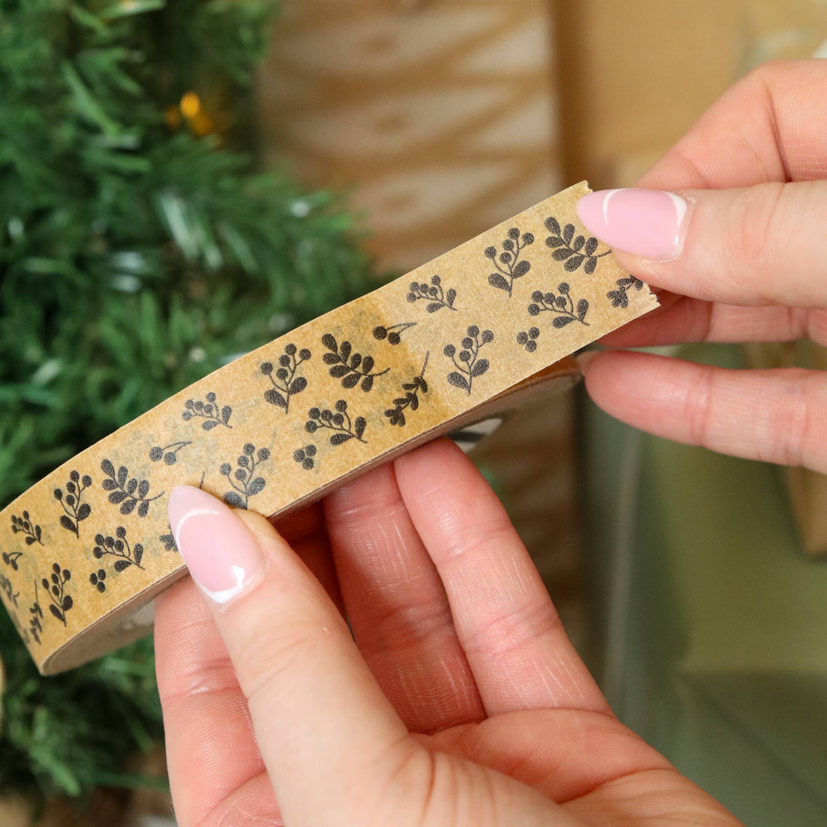 A close-up of hands holding a roll of decorative paper tape with a black botanical pattern on a kraft paper background, with a blurred green pine tree in the background.