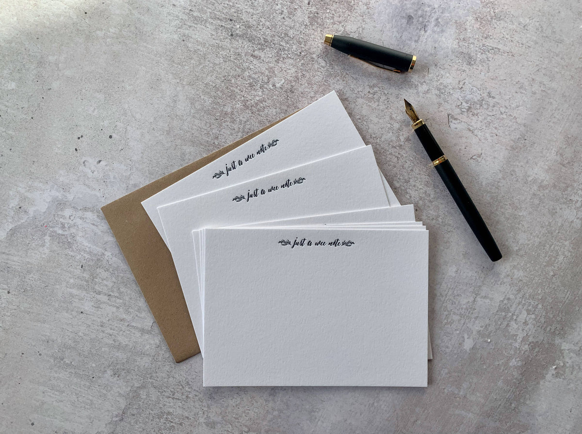 A stack of white stationery cards with black cursive writing, a brown kraft envelope, and two black fountain pens with gold accents, arranged on a textured light gray surface.