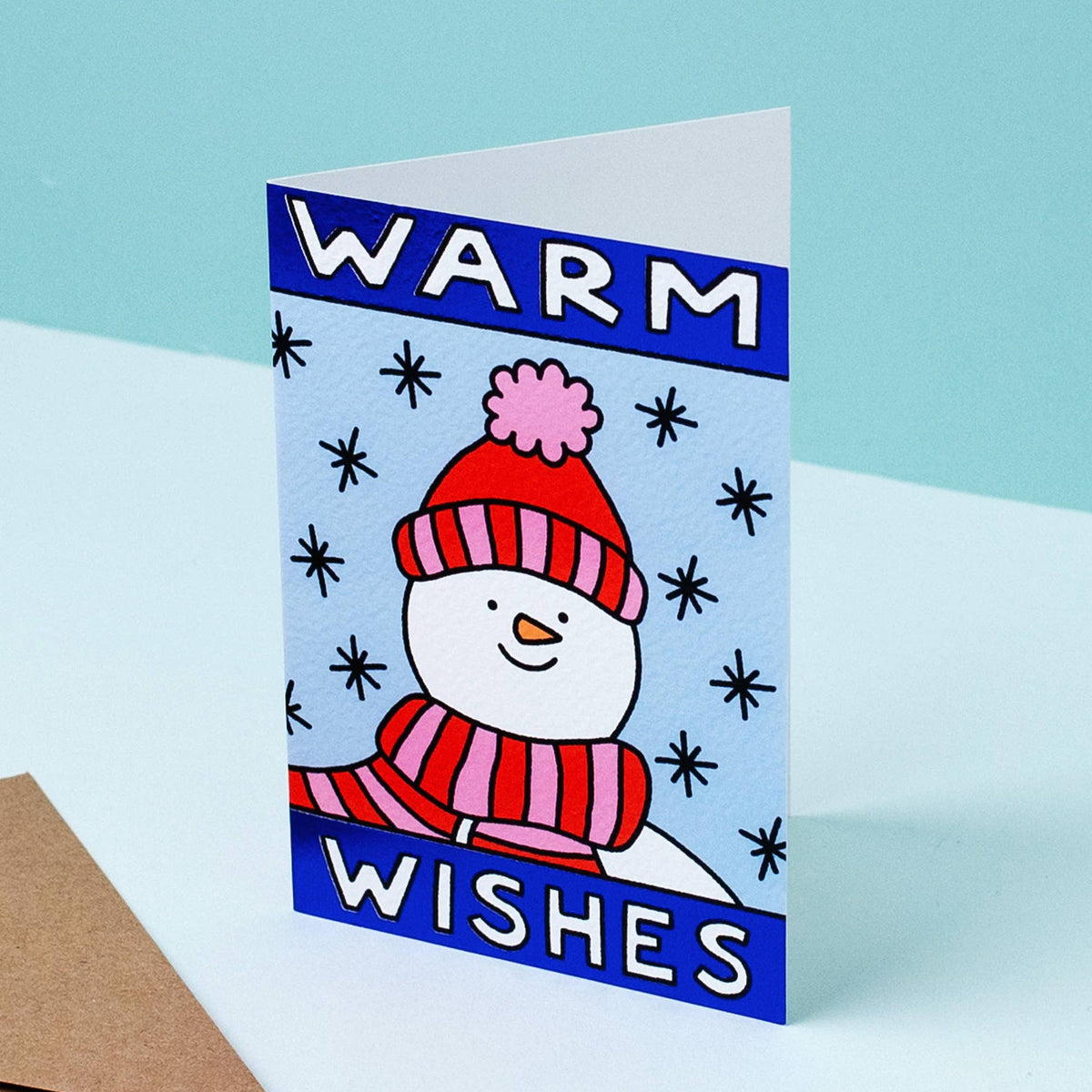 A folded greeting card with a winter illustration of a snowman wearing a red striped hat and scarf, with the words 'WARM WISHES' on a light blue background. A brown envelope sits beside it on a pale surface against a light blue backdrop.