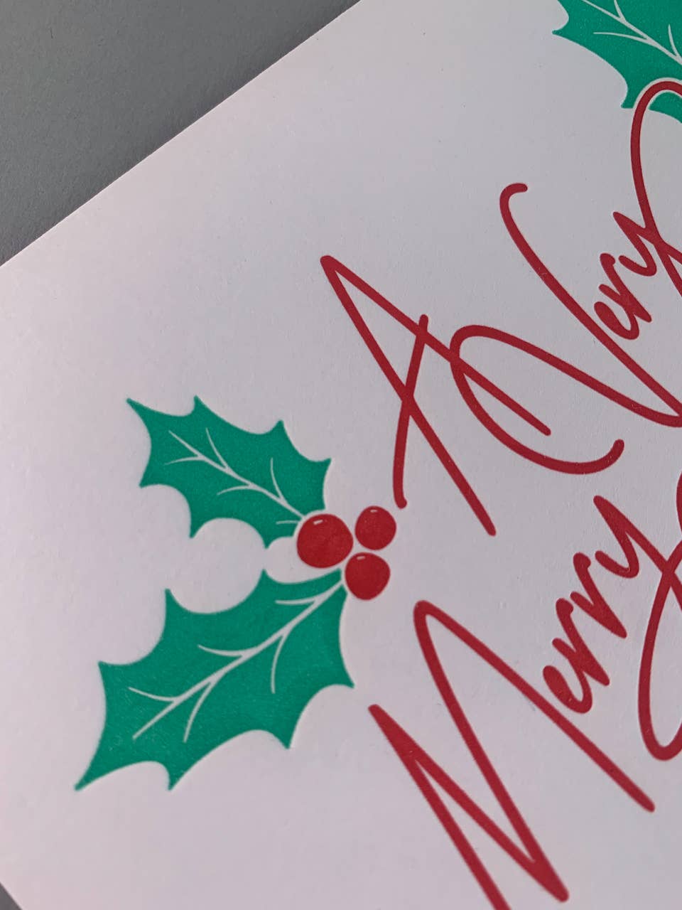 A close-up of a white greeting card featuring green holly leaves with red berries and red cursive holiday text, photographed against a grey background.