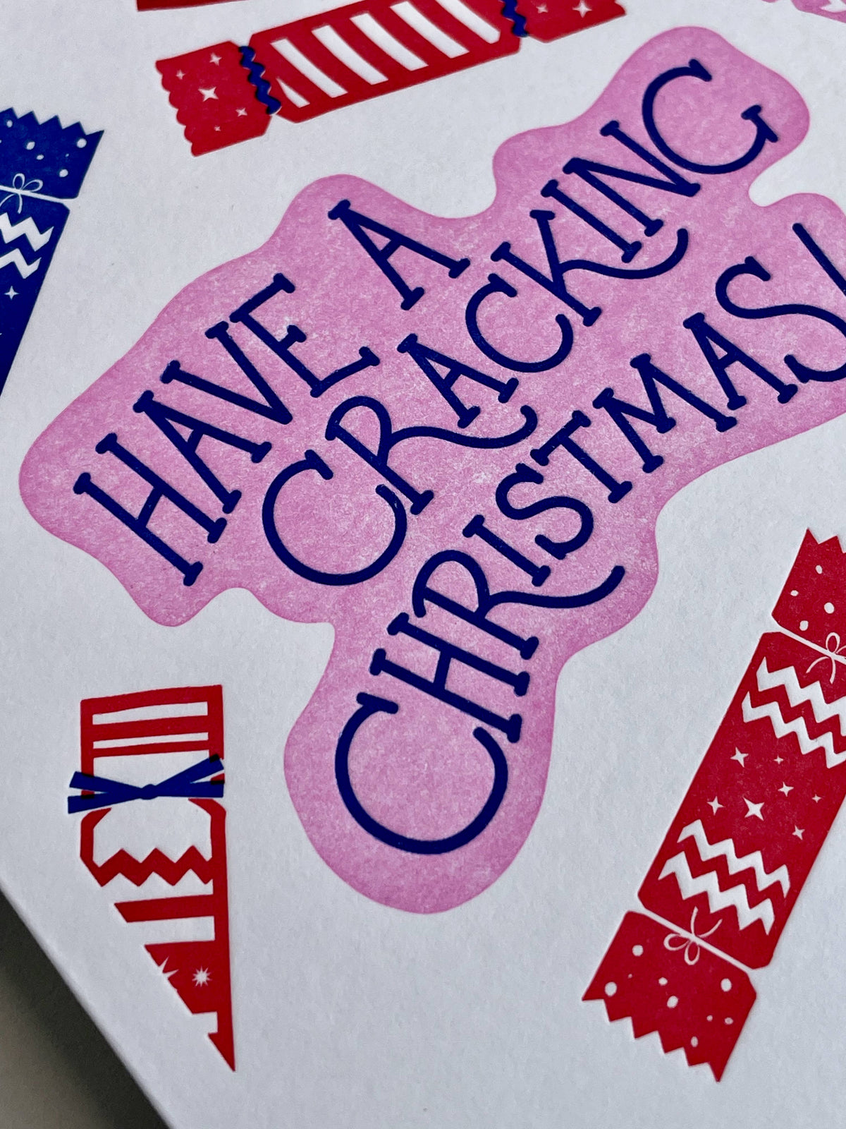 A close-up of a printed christmas design with the phrase “HAVE A CRACKING CHRISTMAS!” in dark blue lettering on a pink shape, surrounded by red and blue Christmas crackers on a white background.