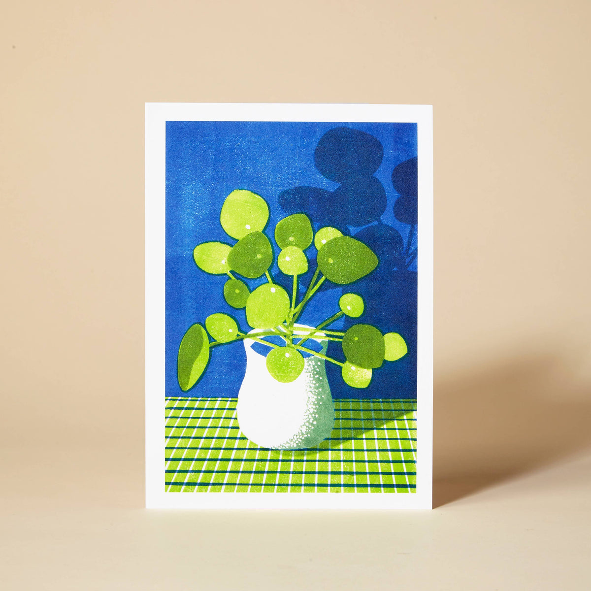 A greetings card with a green pilea money plant in a white vase on a blue background and green tartan tablecloth