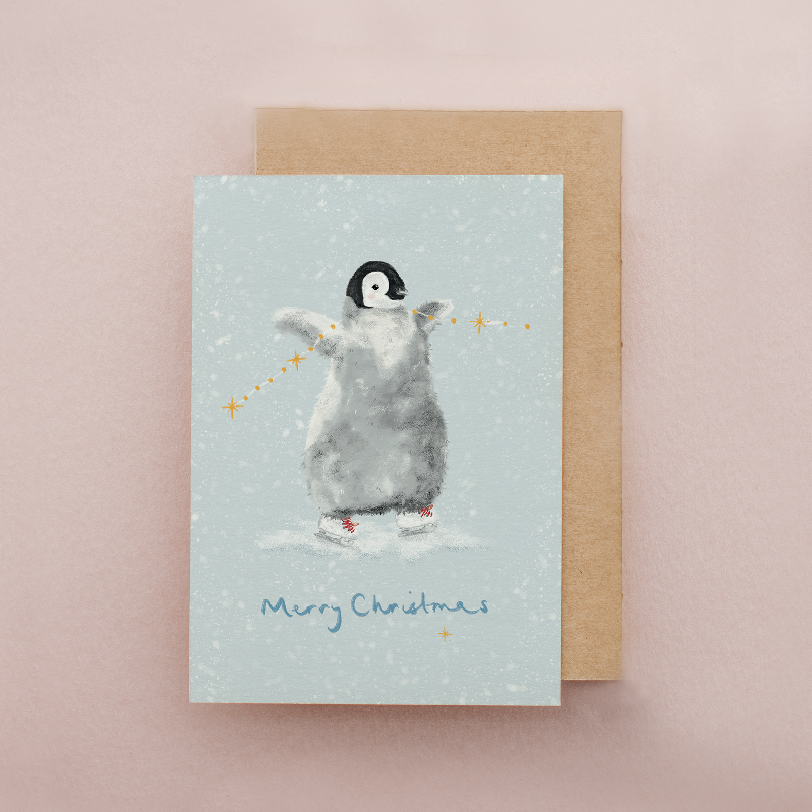A Christmas greeting card featuring a penguin standing on ice with stars around it, placed in front of a brown kraft envelope on a soft pink background.
