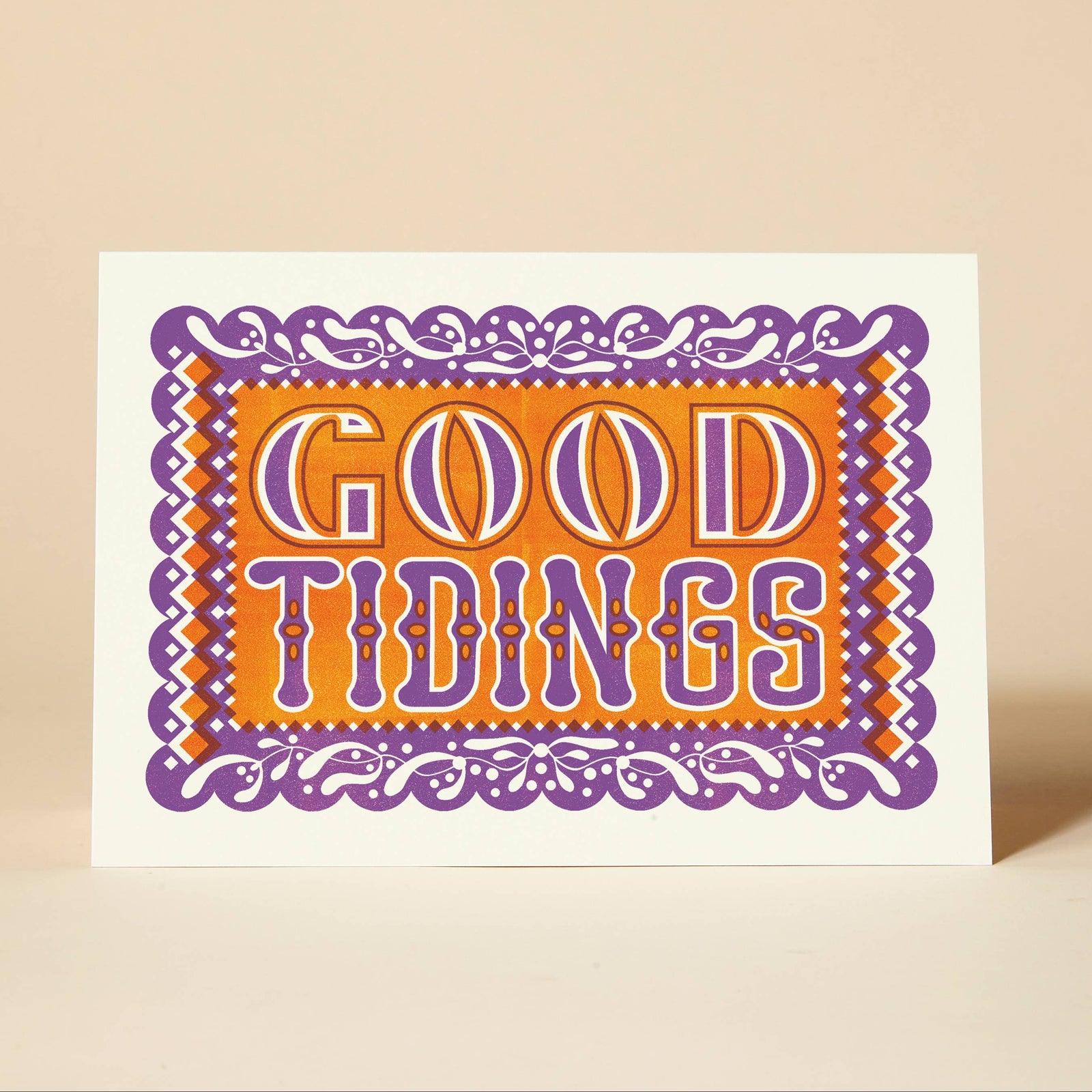 A white Christmas greeting card with a colourful 'GOOD TIDINGS' design in purple and orange, standing upright on a beige surface against a matching beige background.