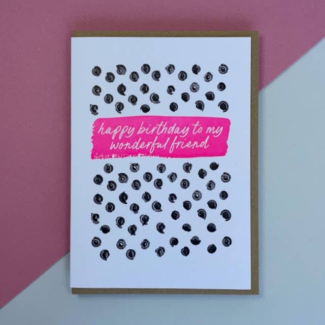 A letterpress printed birthday card with a white front, decorated with black ink dots and a bright pink banner reading 'happy birthday to my wonderful friend'. The card is shown on a brown kraft envelope, set against a diagonally split pink and light blue background.