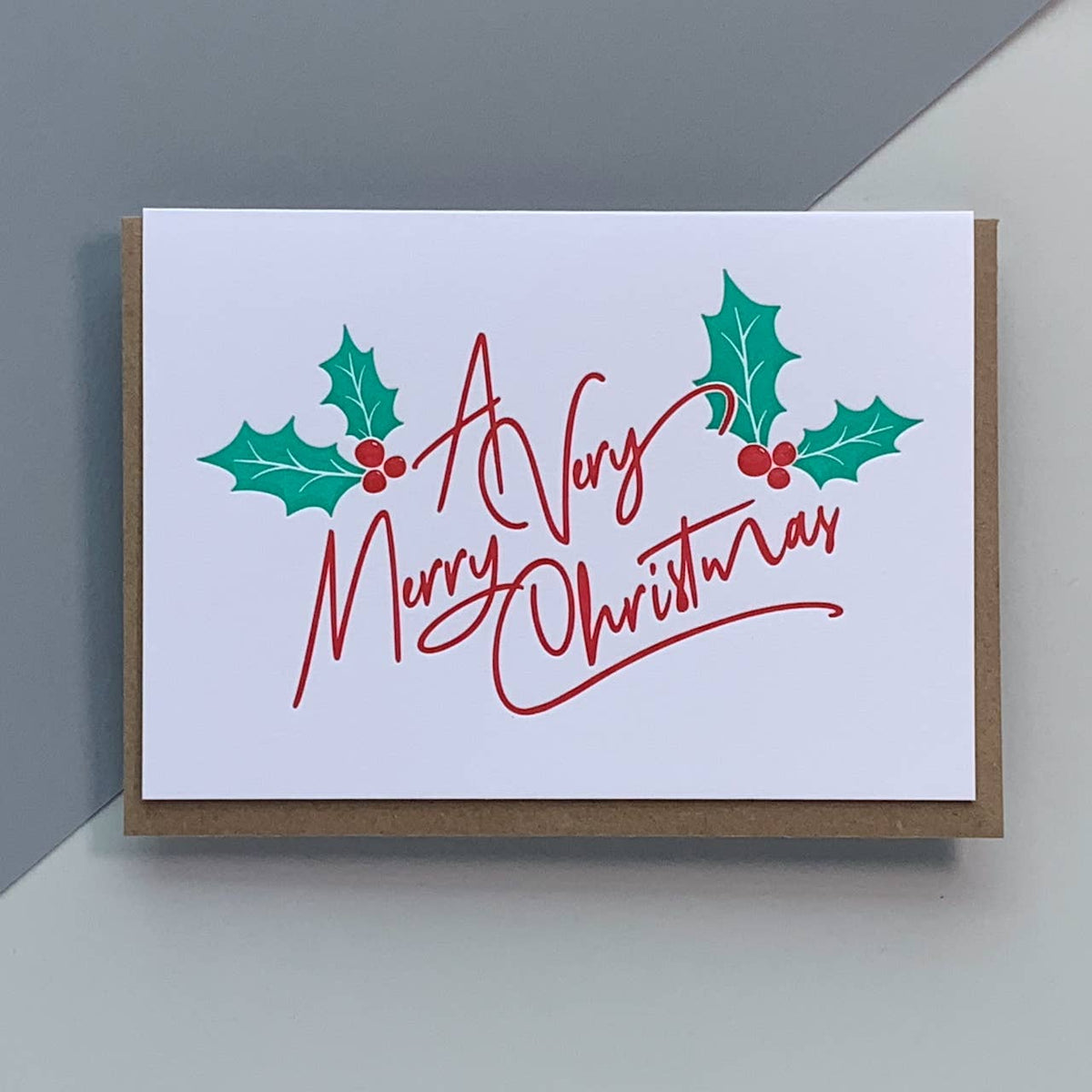 An A6 letterpress Christmas greeting card with a white front, red cursive script reading 'A Very Merry Christmas,' and green holly leaves with red berries, shown with a brown kraft envelope on a light gray background.