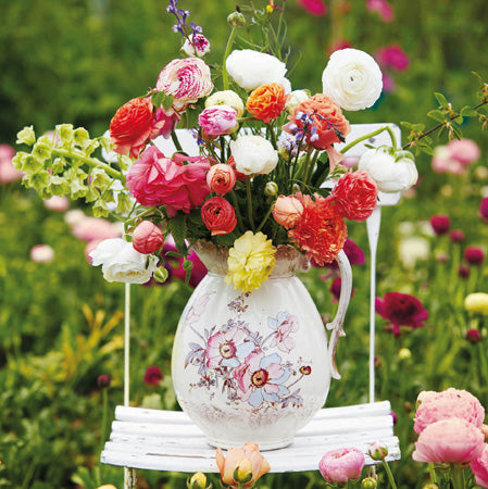 A greetings card with a white floral vintage style vase sitting on a slatted garden chair. The vase is full of a floral arrangement that includes different coloured ranunculus flowers amongst others. The chair sits in a floral garden.