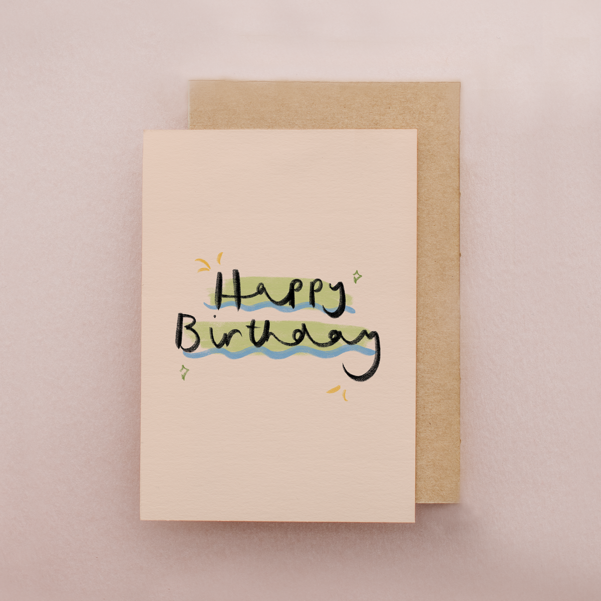 Mini birthday card with multi-coloured 'Happy Birthday' handwritten text on a cream background showing kraft brown envelope behind
