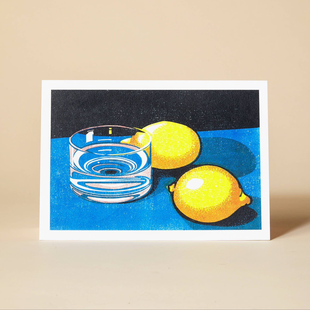 A greetings card featuring a still life illustration of two yellow lemons and a clear glass of water on a blue and black background, displayed on a beige surface.