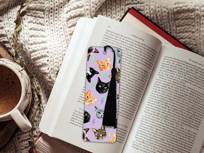 A image of an open book sitting on a knitted blanket and a mug of hot chocolate. The book has inside a long rectangular shaped bookmark with long black tassel. The design on the bookmark has a lilac background and is covered in cutout cat heads of different types of cats - tabby, ginger, black and white, grey.