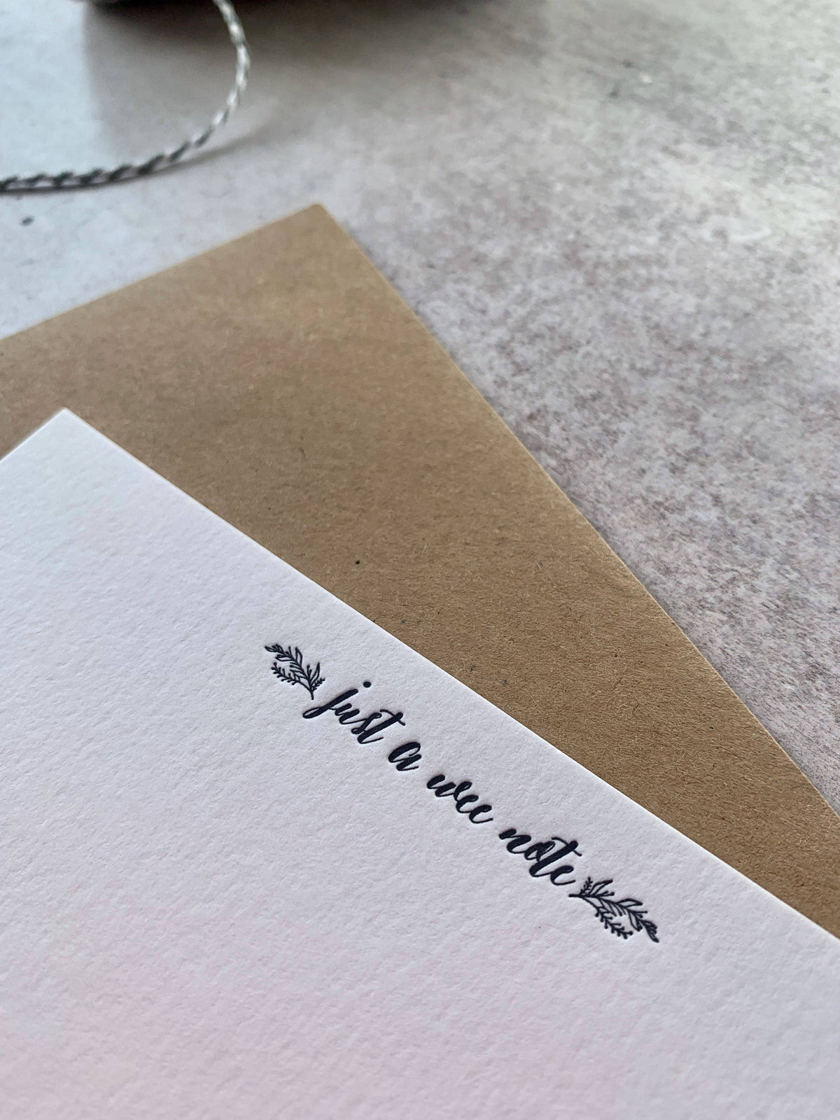 A close-up of a white textured card printed with the phrase 'just a wee note' in black script, layered over a brown kraft envelope on a light stone-like surface.