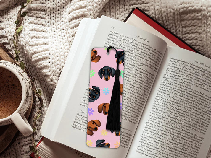 A image of an open book sitting on a knitted blanket and a mug of hot chocolate. The book has inside a long rectangular shaped bookmark with long black tassel. The design on the bookmark has a light pink background and is covered in cutout dog heads of different types of dachshunds- black and tan and brown only. There are holographic flowers inbetween.