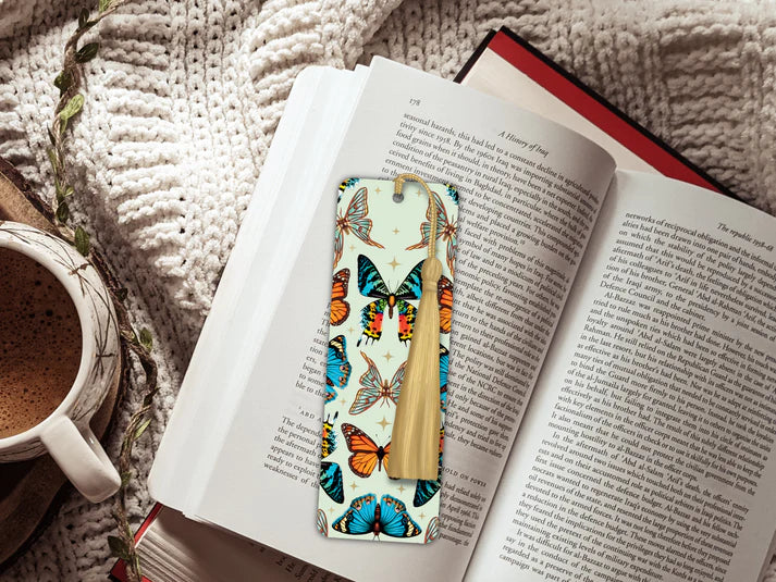 A image of an open book sitting on a knitted blanket and a mug of hot chocolate. The book has inside a long rectangular shaped bookmark with long gold tassel. The design on the bookmark has a light green background and is covered in cutout of different types of butterflies. There are holographic stars inbetween.