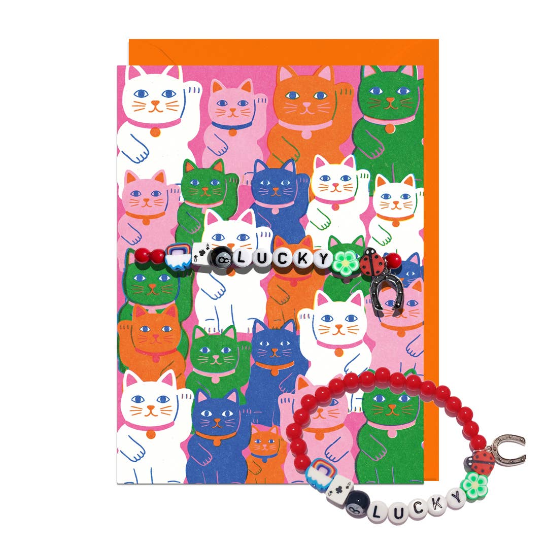 A colourful greeting card with a matching beaded LUCKY letter bracelet. The card has a repeating pattern of illustrated lucky cats in various colours (green, blue, orange, pink, white) on a white background, with an orange envelope behind it.