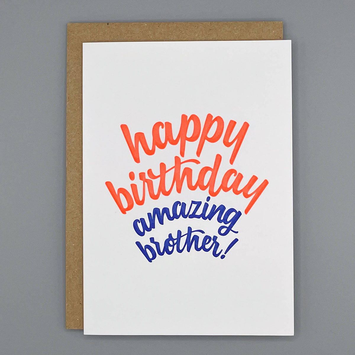 A cream letterpress printed greeting card with the phrase 'happy birthday amazing brother!' in orange and blue script, shown with a brown kraft envelope on a gray background.