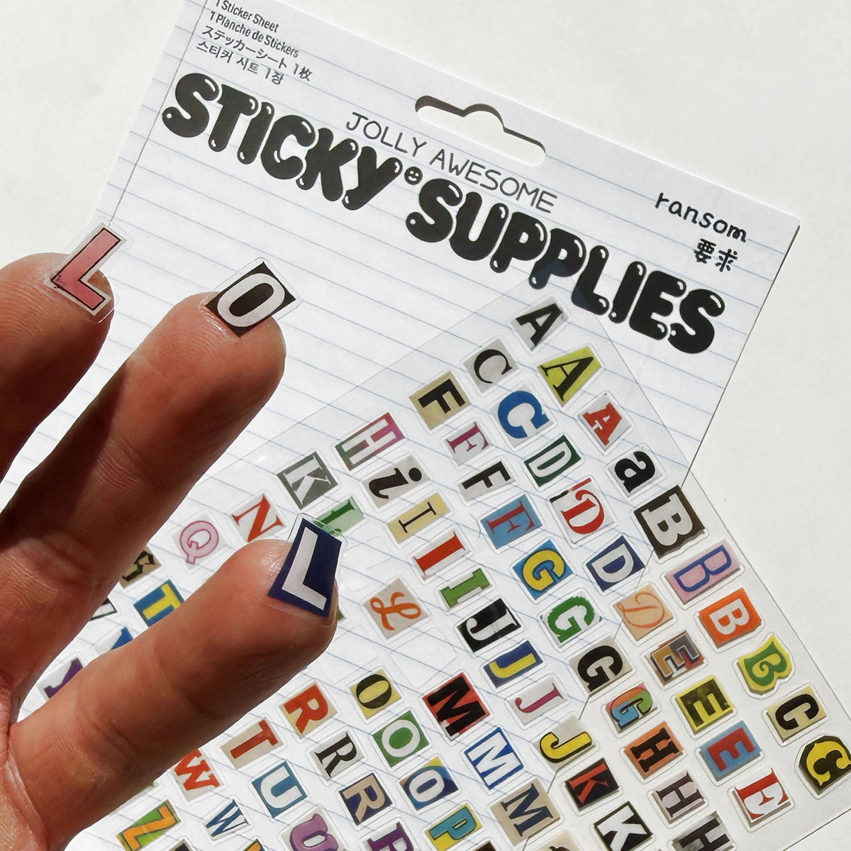 A hand holds a sheet of colourful alphabet random note stickers labelled 'Sticky Supplies' against a white background, with more sticker sheets visible behind it.