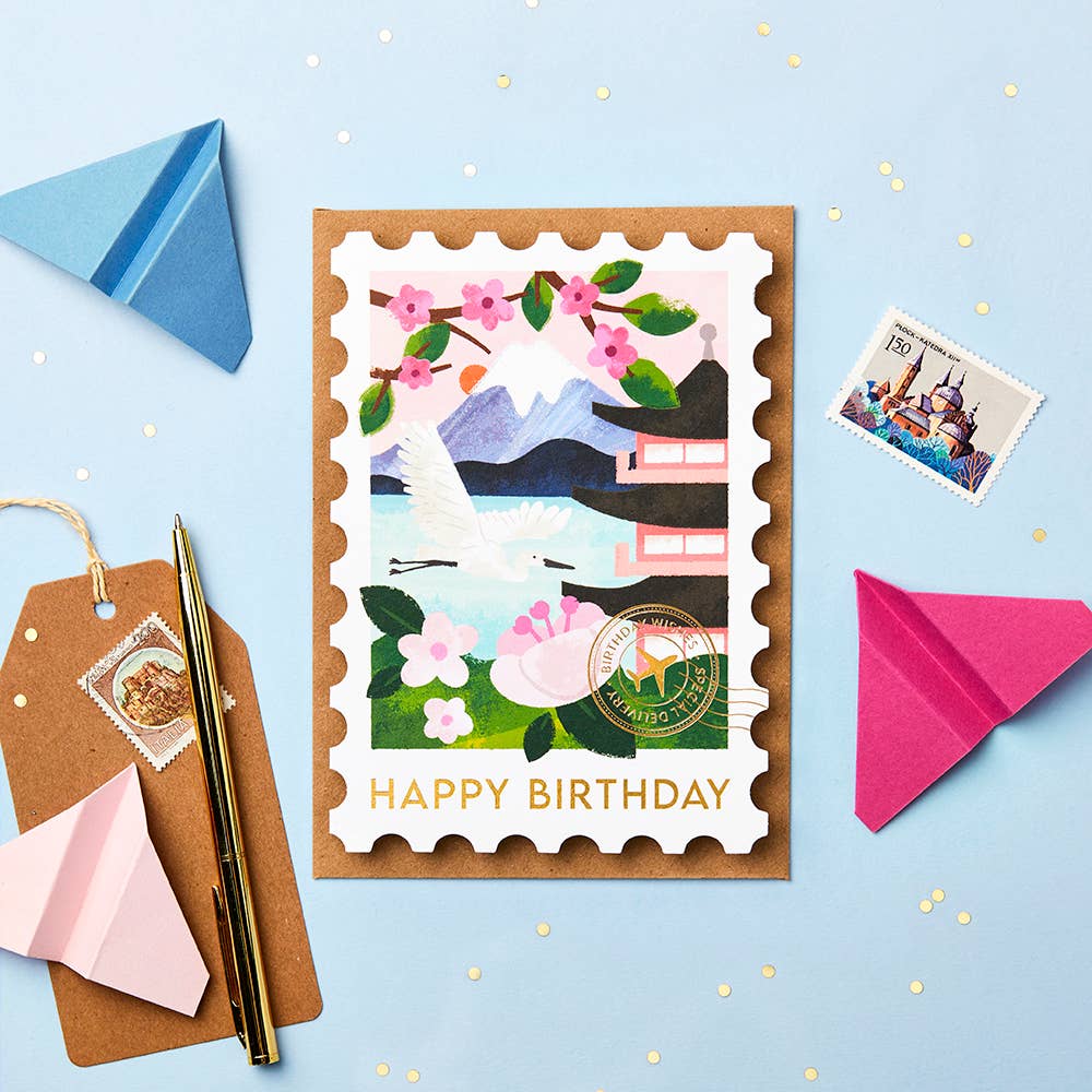 Birthday card with Japanese-themed illustration on a blue background accompanied by a brown envelope and a stamp, paper planes and a luggage tag.