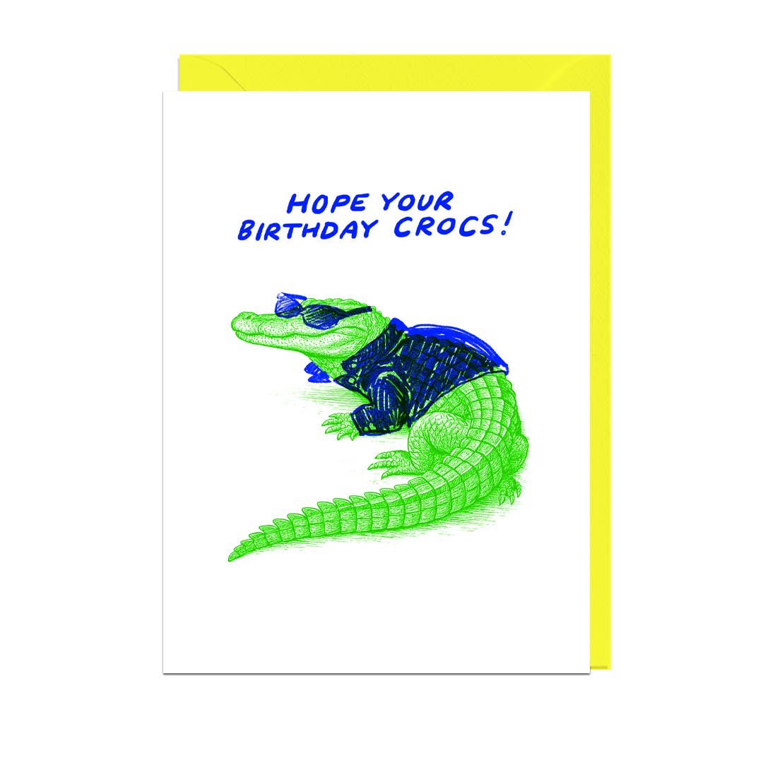 Birthday card with a green crocodile wearing a blue leather jacket and text 'Hope your birthday crocs!' on a white background with yellow envelope.