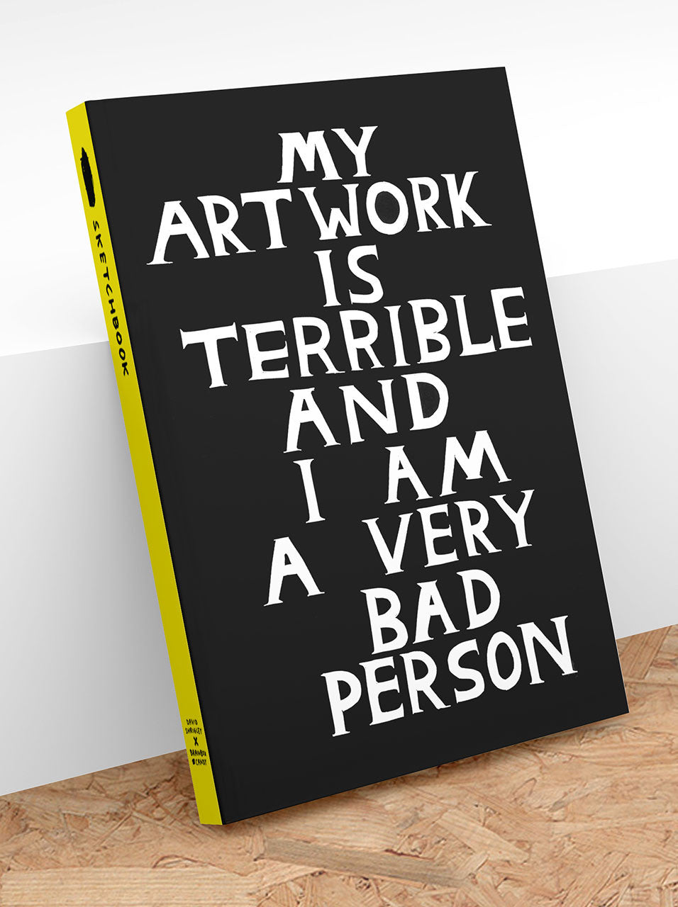Sketchbook with white text 'My artwork is terrible and I am a very bad person' hand written in block capitals on a black background with a yellow binding leaning against a wall.