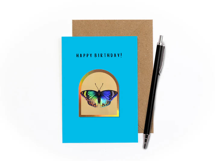 An image of a greetings card with kraft brown envelope tucked into it and a pen sat at an angle to the right. The card has an azure blue background. It features a holographic arch sticker in the centre with a cutout of a butterfly on a beige background.