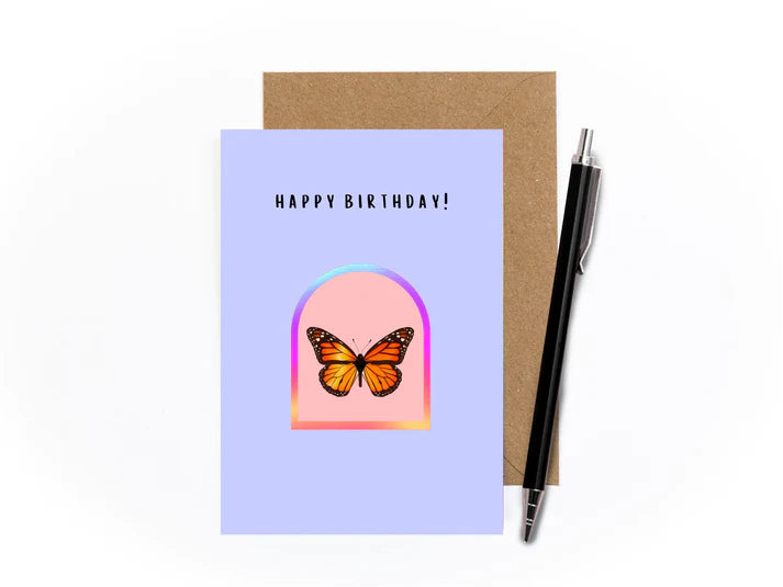 An image of a greetings card with kraft brown envelope tucked into it and a pen sat at an angle to the right. The card has a lilac background. It features a holographic arch sticker in the centre with a cutout of a monarch butterfly on a light pink background.