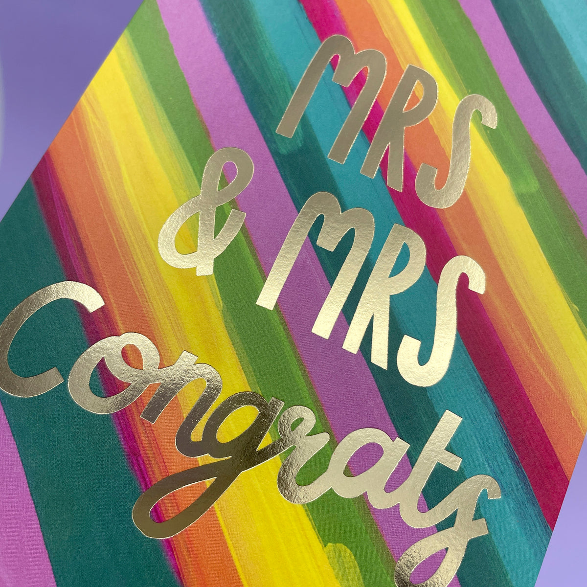 A colourful rainbow stripe greetings card to celebrate a wedding. The rainbow stripes are diagonal across the card and the writing in the centre of the card is big gold foil stating &#39;Mrs &amp; Mrs Congrats&#39;.