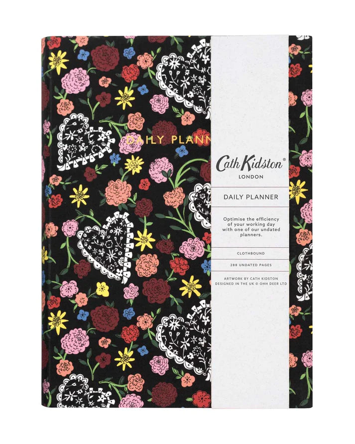 The front cover of a daily planner by Cath Kidston. The cover design has a black background and is covered in colourful flowers  in burgundy, peach, yellow and pink. It also has lacy hearts dotted through the design. There are gold embellished words saying in capitals 'DAILY PLANNER'. The image shows a white belly band including details about the product.