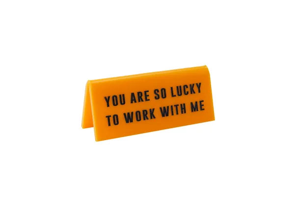 A small yellow acrylic sign that has in big black capital letters on it &#39;YOU ARE SO LUCKY TO WORK WITH ME&#39;.