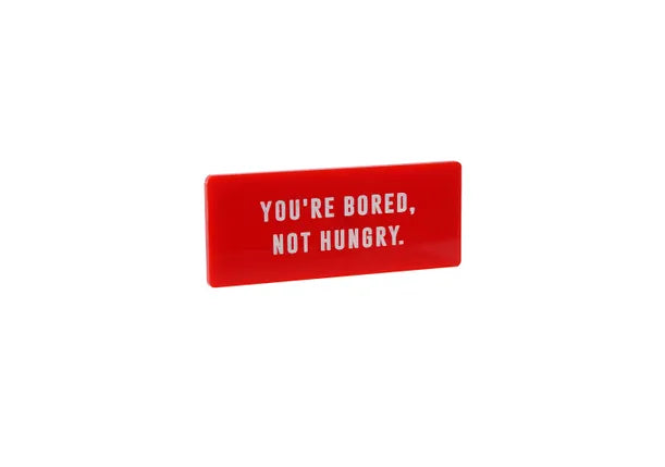 A rectangular red fridge magnet with the words YOU'RE BORED NOT HUNGRY in white capitals.