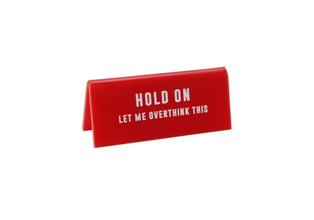 A small red acrylic sign that has in big white capital letters on it &#39;HOLD ON LET ME OVERTHINK THIS&#39;.