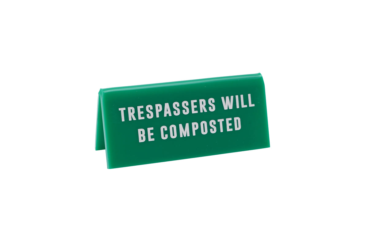 A small GREEN acrylic sign that has in big white capital letters on it &#39;TRESSPASSERS WILL BE COMPOSTED&#39;.