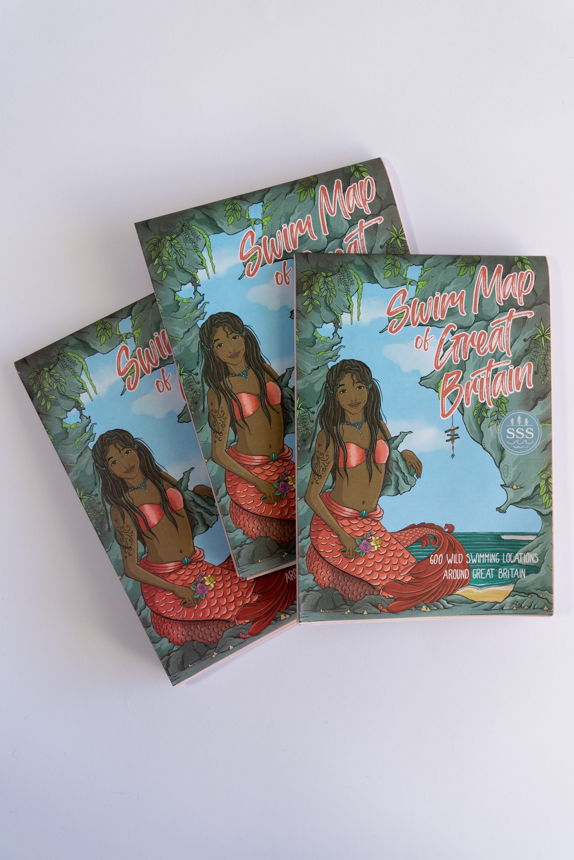 Three front covers of a swim map of Great Britain showing a mermaid at the entrance to a cave. The mermaid has long dark brown hair, black skin colour and a red tail.