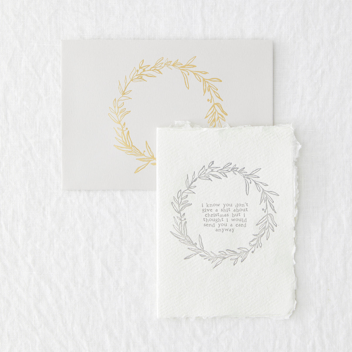 An image of a letterpressed christmas greetings card and envelope. The light grey envelope has a ring of gold foliage in the centre and the card has teh same but printed in grey with the words in the centre &#39;I know you don&#39;t give a shit about christmas but I thought I would send you a card anyway&#39;.