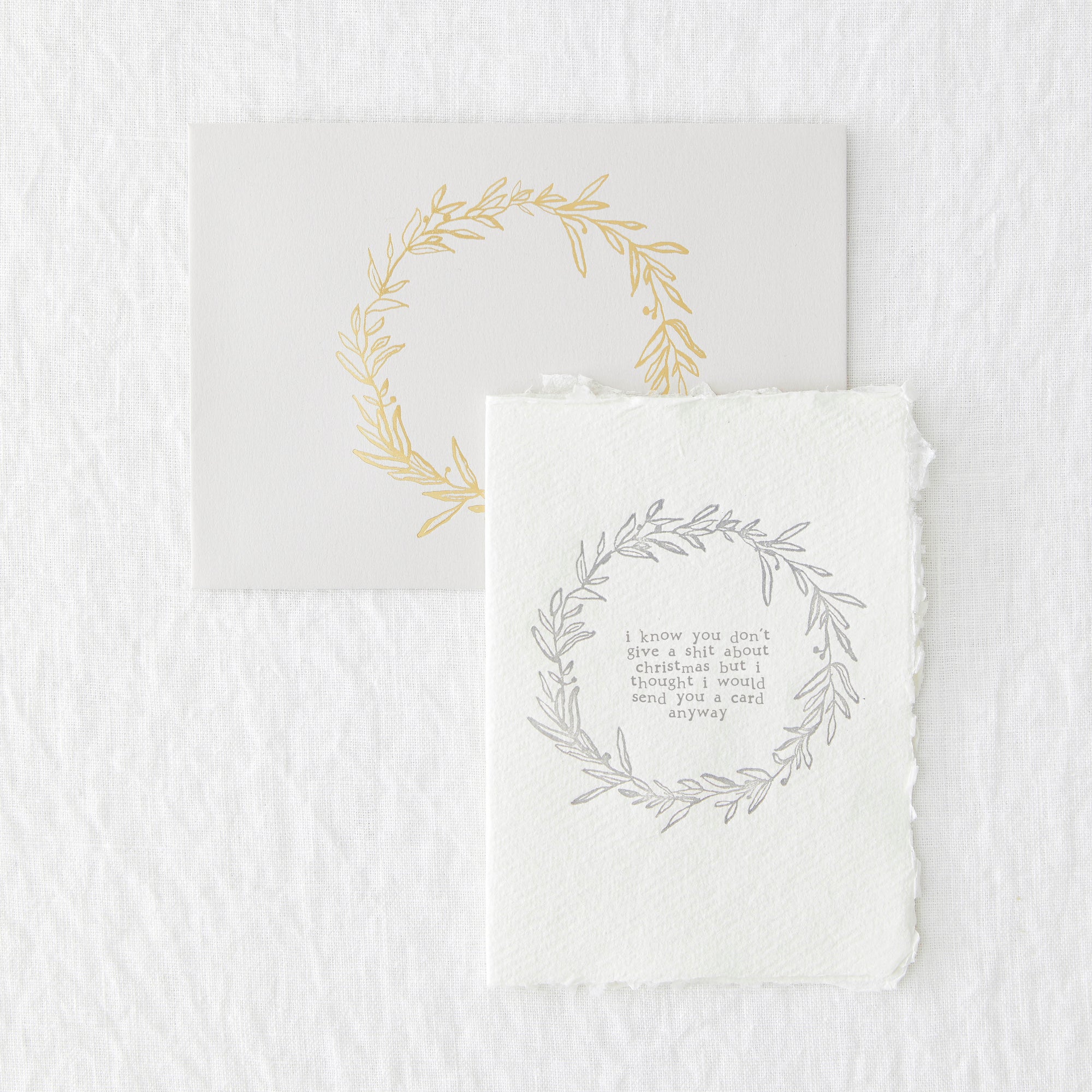 An image of a letterpressed christmas greetings card and envelope. The light grey envelope has a ring of gold foliage in the centre and the card has teh same but printed in grey with the words in the centre 'I know you don't give a shit about christmas but I thought I would send you a card anyway'.
