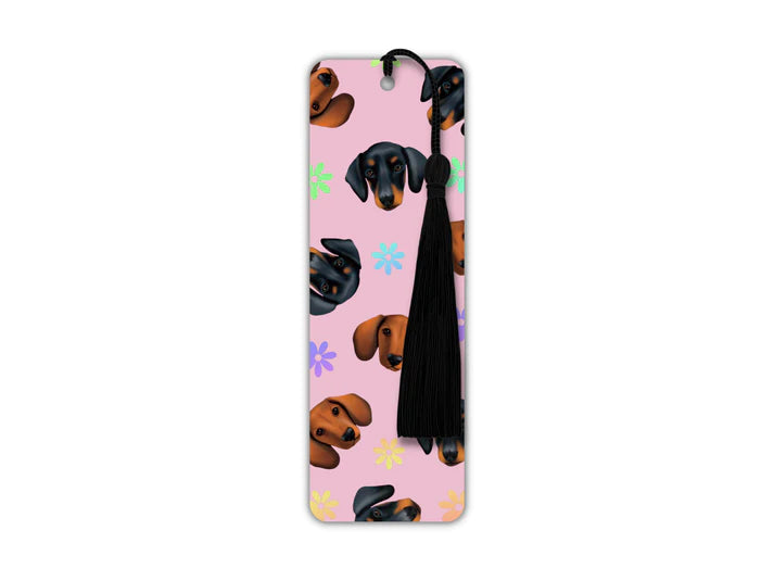 A long rectangular shaped bookmark with long black tassel. The design on the bookmark has a light pink background and is covered in cutout dog heads of different types of dachshunds- black and tan and brown only. There are holographic flowers inbetween.